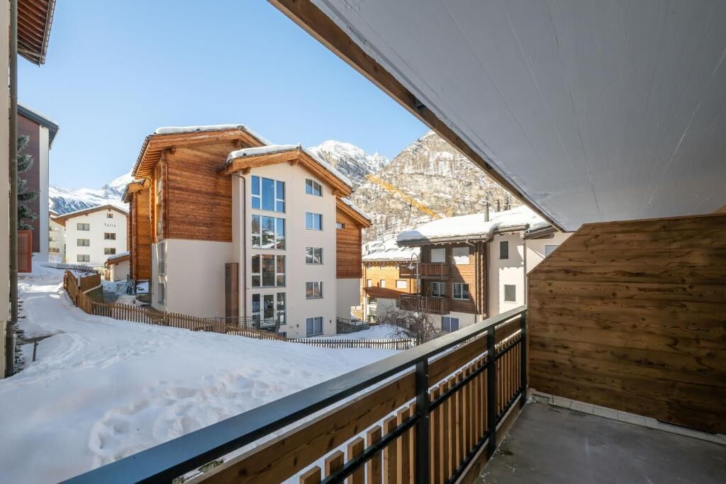 A snow-covered balcony with a wooden railing overlooking a residential area with buildings and mountains in the background.