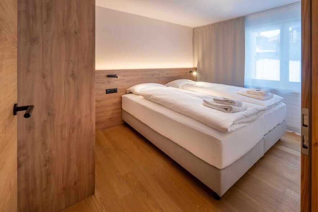 A modern hotel room with a wooden floor, a large bed with white linens, a bedside table with a lamp, and a window with curtains.