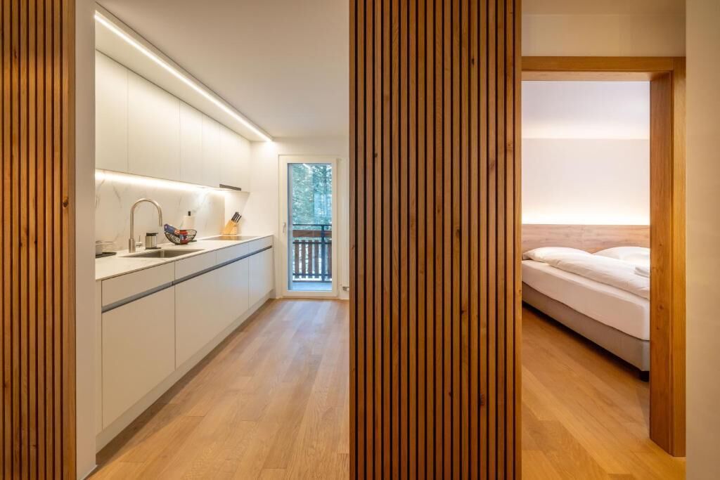 Una cocina moderna con armarios blancos y piso de madera, que da paso a un dormitorio con cama y balcón.