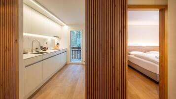 Una cocina moderna con armarios blancos y piso de madera, que da paso a un dormitorio con cama y balcón.
