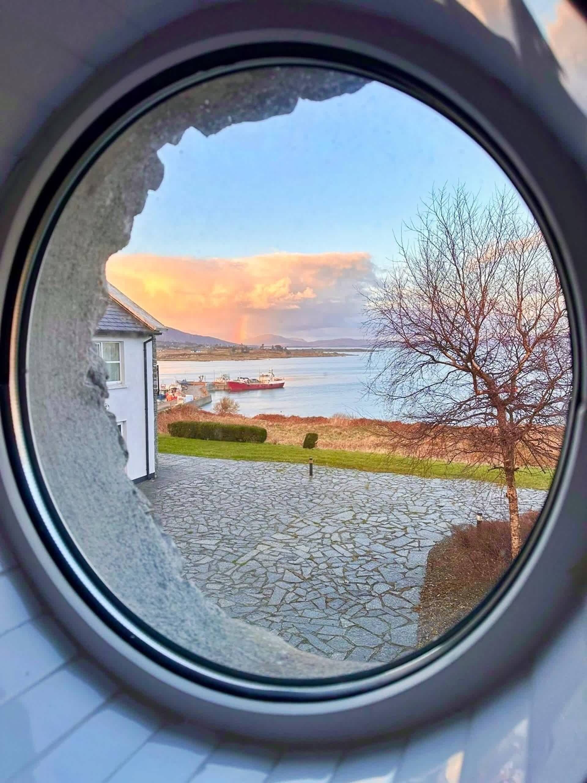A view through a circular window of a riverside area with a small building and a tree.