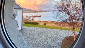 A view through a circular window of a building with a stone-paved area, a small white structure, and a tree. - Oyster Cove Kilkerrin stunning views (Kilkerrin)