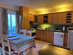 A kitchen with wooden cabinets, a dining table with chairs, a window with curtains, and a green-tiled backsplash. - Oyster Cove Kilkerrin with stunning views (Kilkerrin)