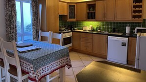 A kitchen with wooden cabinets, a dining table with chairs, a stove, a dishwasher, and a window with curtains. - Oyster Cove Kilkerrin stunning views (Kilkerrin)