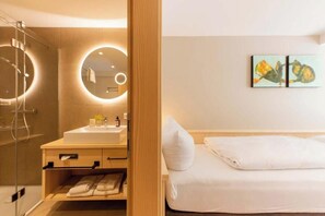 A hotel room with a bathroom featuring a sink, mirror, and towel rack. There is a bed with white bedding and a painting on the wall.