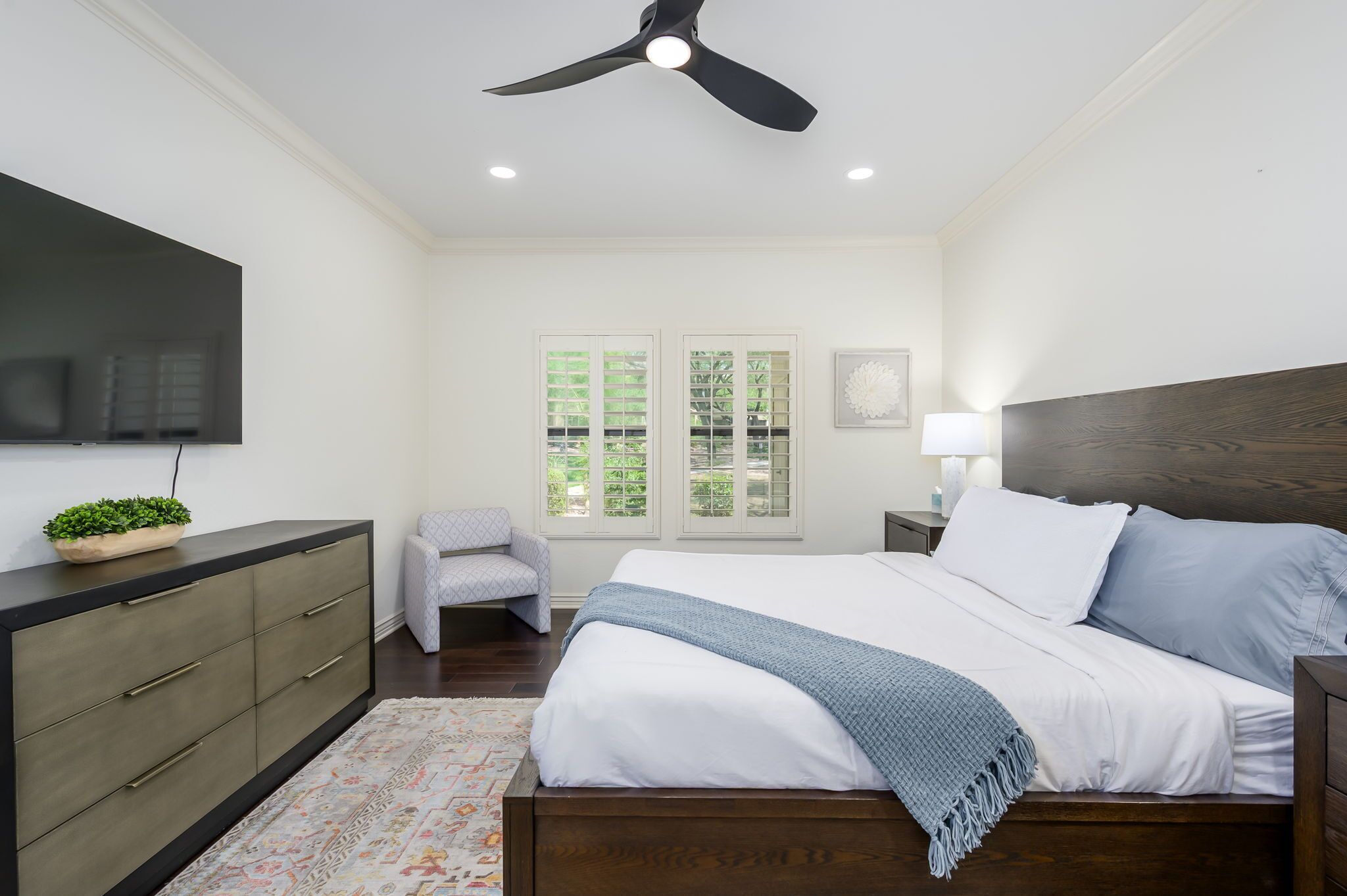 A bedroom with a bed, a TV, a ceiling fan, a window with blinds, a nightstand, a lamp, and a dresser.