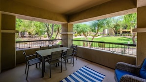 A covered patio with a table and chairs, a blue and white striped rug, and a view of a well-maintained garden and building. - Highly desired Desert Ridge condo on 1st level- no stairs, large patio (Phoenix)