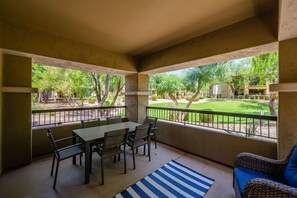 Um alpendre coberto com uma mesa e cadeiras, um tapete listrado em azul e branco, e vista para um jardim bem cuidado e um edifício.