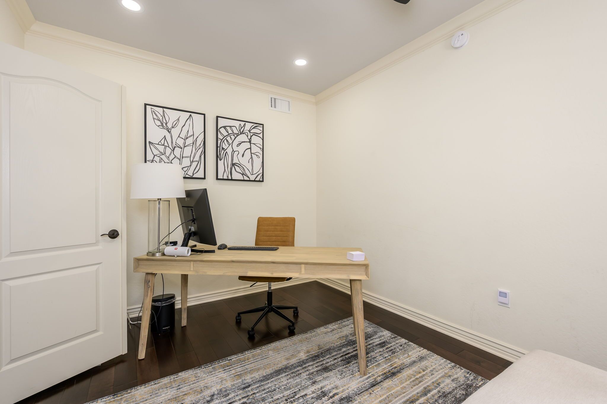 Een kamer met een bureau, stoel, lamp en twee ingelijste kunstwerken aan de muur.
