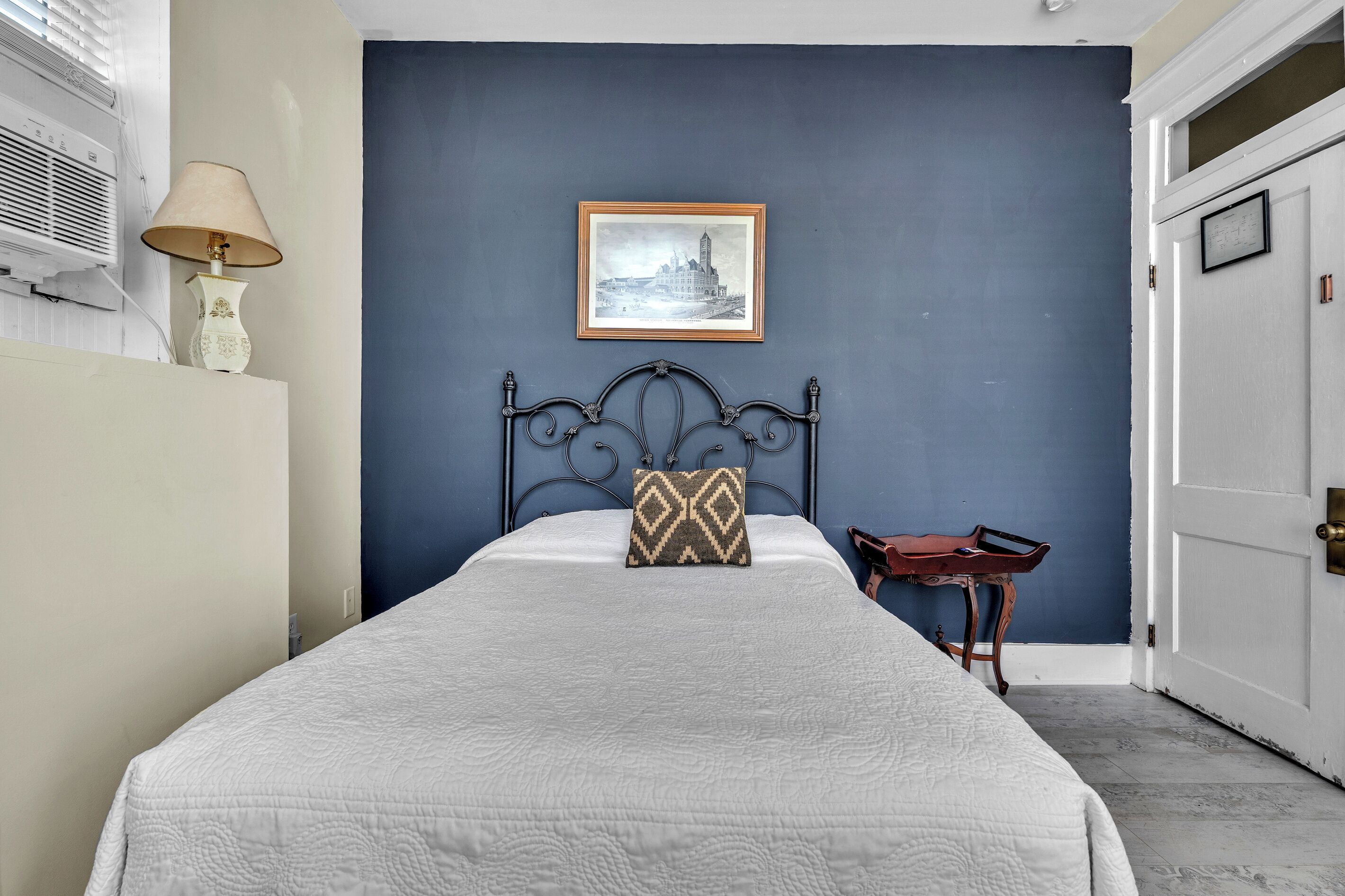 A bedroom with a bed, a framed picture on the wall, a bedside table, and a lamp.