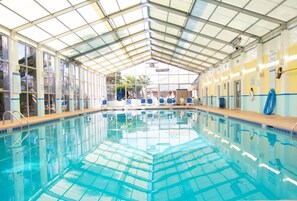 Une piscine intérieure avec des parois en verre transparent, un sol carrelé et un toit soutenu par des poutres métalliques.