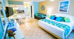 Quarto de hotel com cama, escrivaninha, poltrona, televisão, sofá e cozinha.