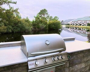Una parrilla de acero inoxidable con un termómetro digital, frente a una pared de piedra con vistas a un cuerpo de agua y edificios al fondo.