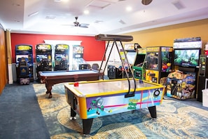 Una mesa de billar con diseños coloridos, juegos de arcade y un ventilador de techo en una sala con alfombra estampada.