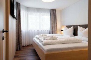 Ein Hotelzimmer mit Bett, Nachttischlampe, einem Fenster mit Vorhängen und einem an der Wand befestigten Fernseher.