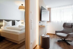 A modern hotel room with a bed, a television mounted on the wall, a seating area with a chair, and a small round ottoman. - Apartment Zamangspitze 32 (Gaschurn)