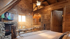 A cozy wooden cabin room with a stone fireplace, a bed with a headboard, a television mounted on the wall, and a ceiling fan. - Smoky Mtn Views  Hot Tub  Sauna Game Room (Sevierville)
