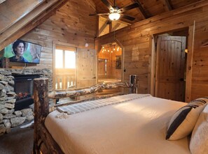 Ein gemütliches Holzblockhaus-Zimmer mit Kamin aus Stein, einem Bett mit Kopfteil, einem an der Wand montierten Fernseher und einem Deckenventilator.