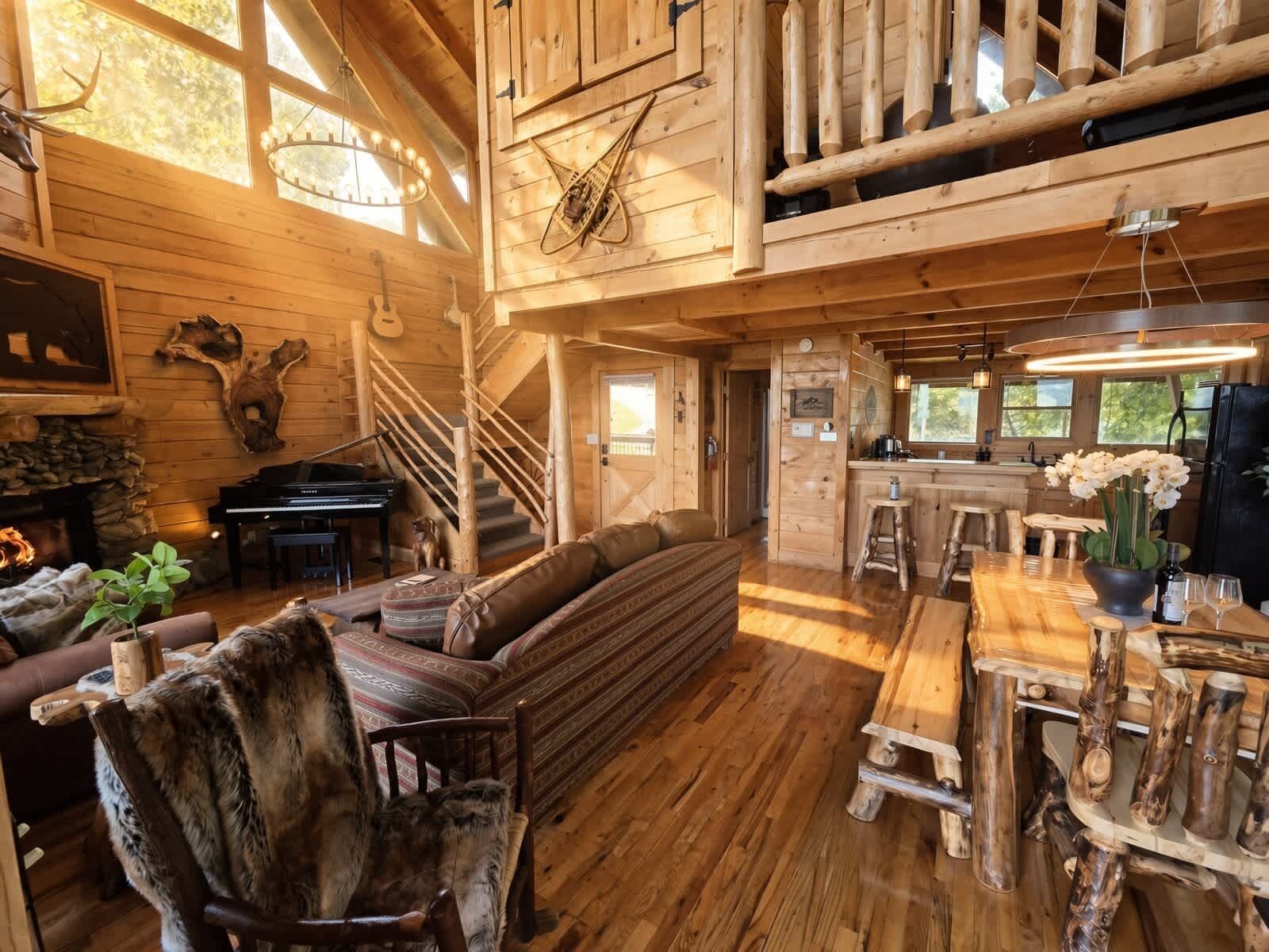 A log cabin interior with a grand staircase, a piano, a dining area with a wooden table, and a fireplace.