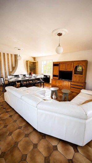 A living room with a large white sectional sofa, a dining area with a table set for four, and a kitchen area with wooden cabinets and a TV. - Logement 100m² - La Villa' Tyno - 9 couchages (La Teste-de-Buch)