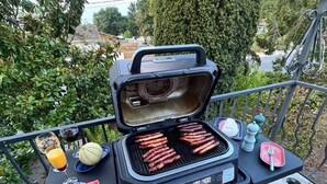 A portable grill with sausages cooking, a watermelon slice, a glass of orange juice, and a bottle of water on a balcony table. - Logement 100m² - La Villa' Tyno - 9 couchages (La Teste-de-Buch)