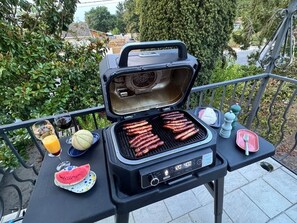 Ein tragbarer Grill mit Bratwürsten, eine Wassermelonenstük, ein Glas Orangensaft und eine Wasserflasche auf einem Balkontisch.
