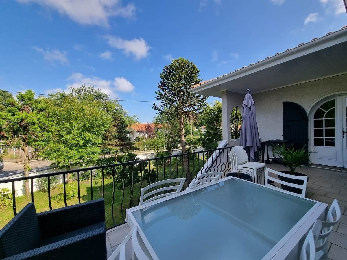 Uma varanda com mesa de vidro e cadeiras brancas, com vista para um jardim e um corpo de água.
