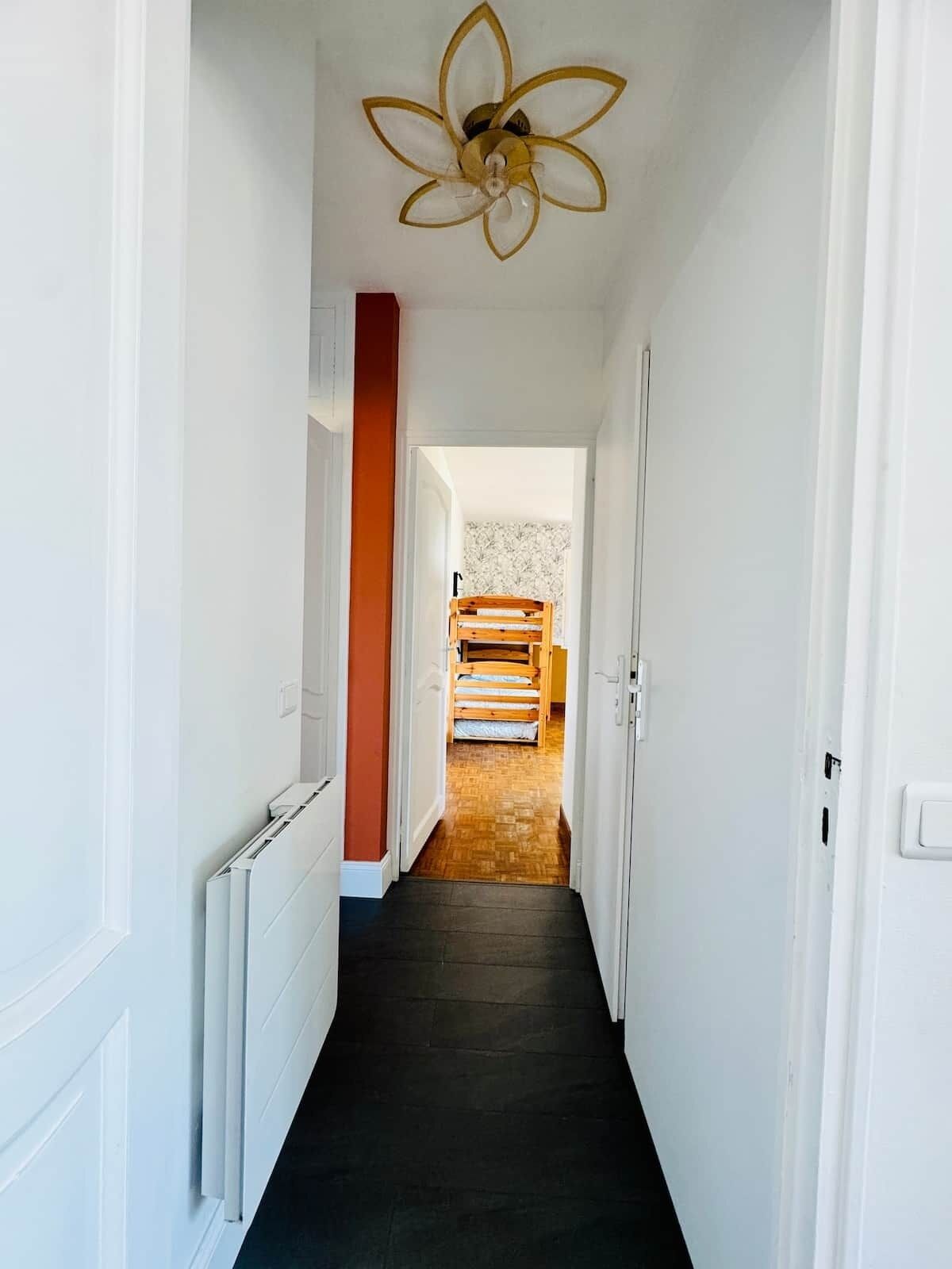 Un couloir étroit avec un radiateur sur le côté gauche, un lit en bois à l’arrière-plan et un ventilateur de plafond orné de motifs floraux.