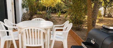 En udendørs terrasse med et rundt bord og hvide plaststole, en grill og en hvid bygning med en sort dør.