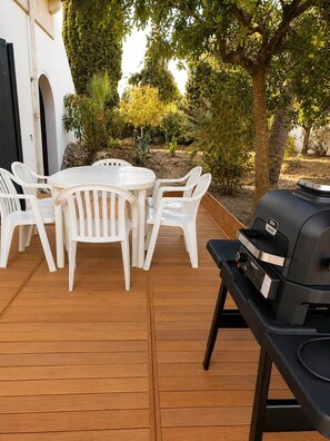 An outdoor patio with a round table and white plastic chairs, a barbecue grill, and a white building with a black door. - Logement RDC - La Villa Tyno Evasion - 60m² (La Teste-de-Buch)