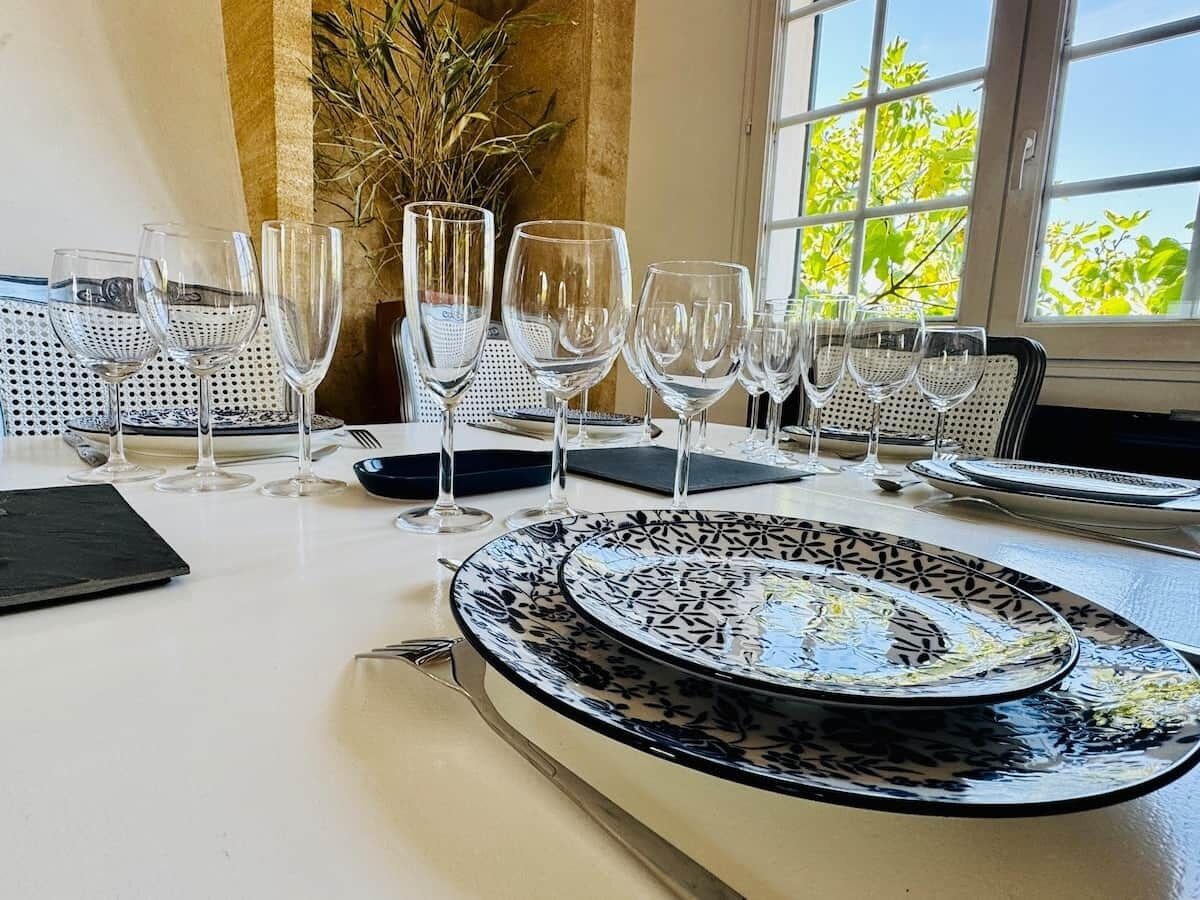 Una mesa de comedor dispuesta con varias copas de vino y platos, un jarrón con plantas secas, y una ventana con vistas a árboles.