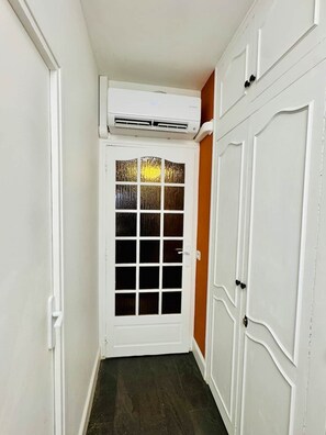 A narrow hallway with a white door featuring a frosted glass panel, a white air conditioning unit above it, and a white wardrobe with a curved design on the right.