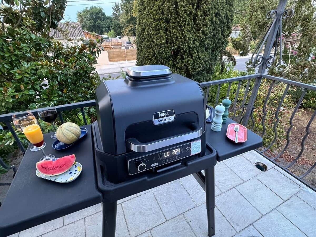 Un barbecue portable doté d’un écran numérique, une tranche de pastèque, un verre de jus d’orange et une bouteille d’eau, le tout posé sur une table à l’extérieur.