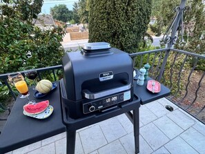 Una parrilla portátil con pantalla digital, una tajada de sandía, un vaso de jugo de naranja y una botella de agua sobre una mesa al aire libre.