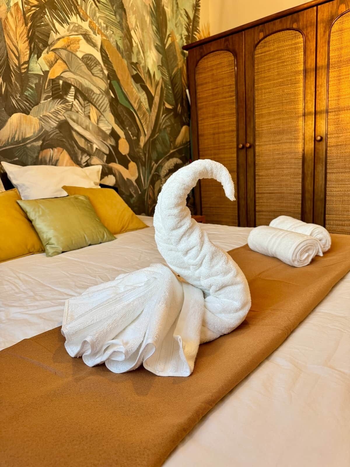 A bed with a swan-shaped towel arrangement, a wooden wardrobe, and a tropical wallpaper.