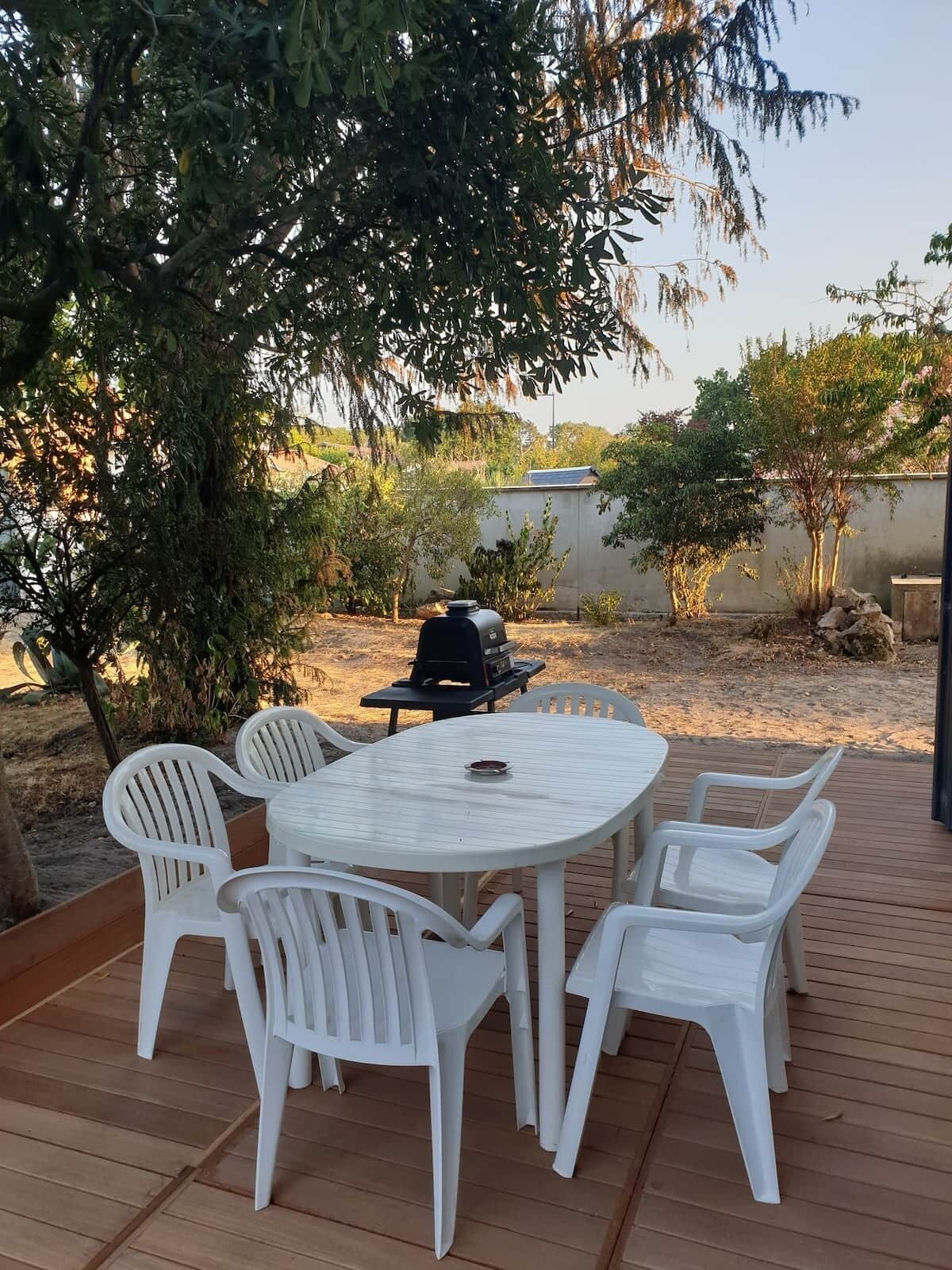 An outdoor patio with a round table and white plastic chairs, surrounded by trees and a barbecue grill.