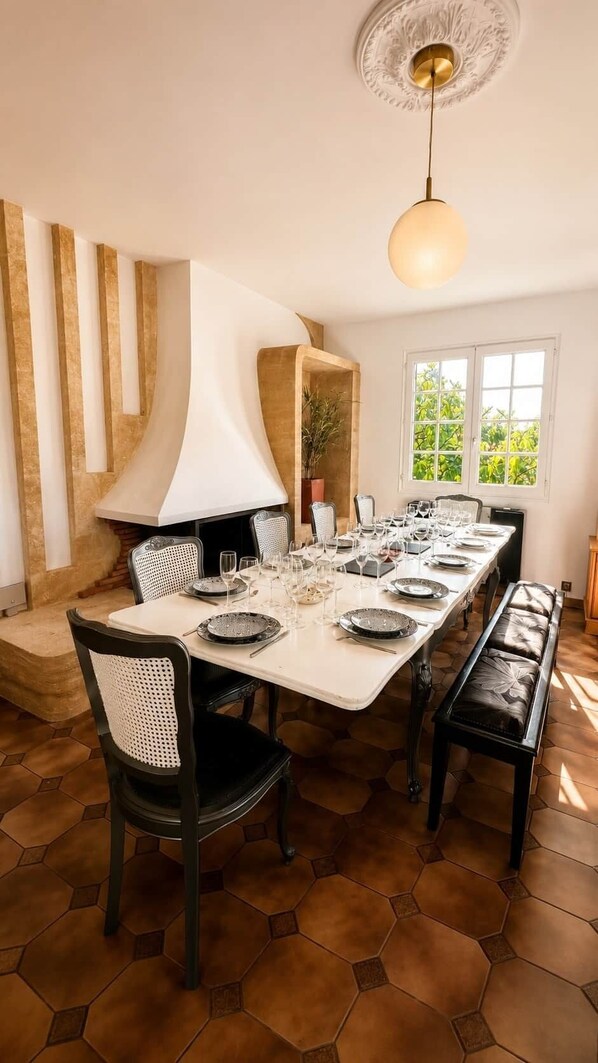 A dining area with a long table set for a meal, chairs, a decorative ceiling light, and a window with a view of greenery.