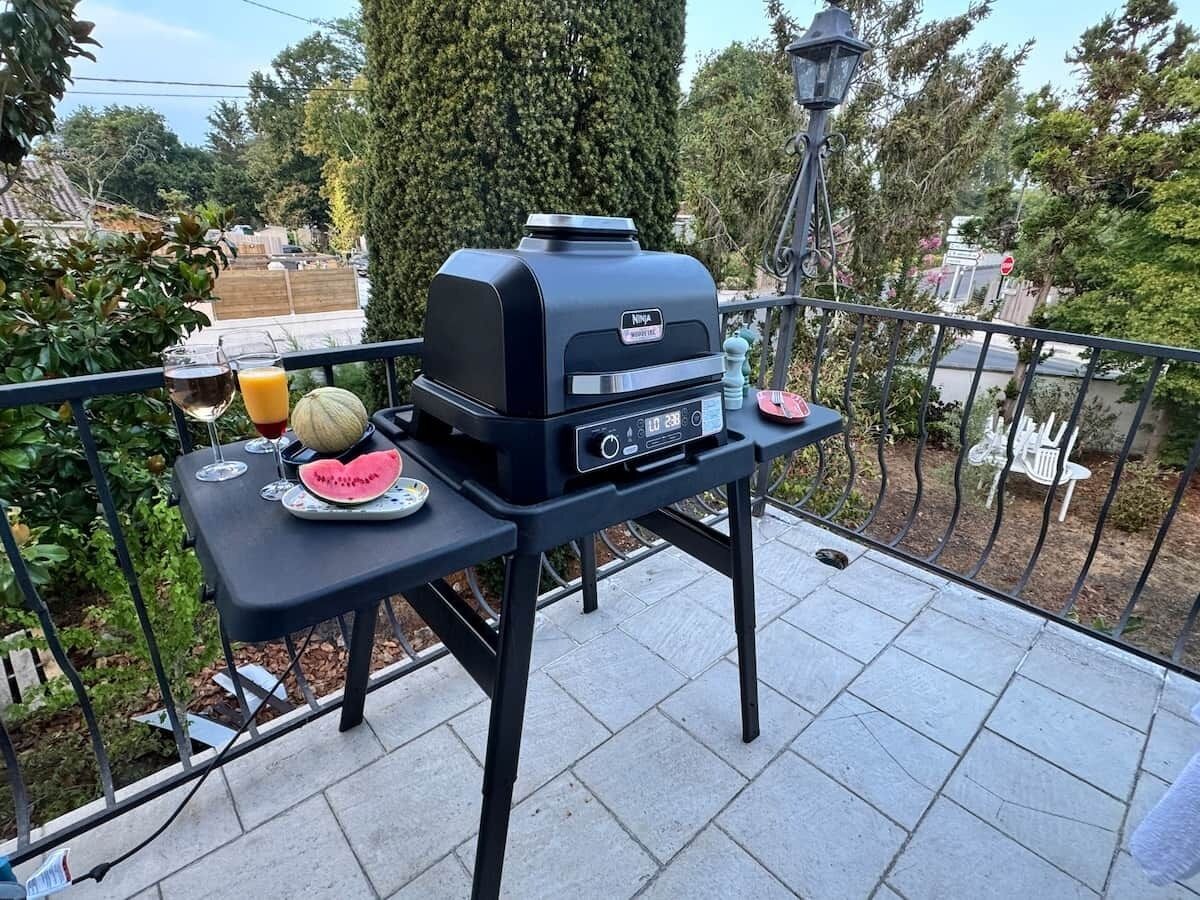A barbecue grill on a balcony with a table, a watermelon, and a glass of orange juice.