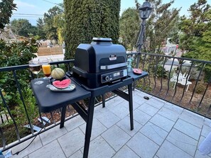 A barbecue grill on a balcony with a table, a watermelon, and a glass of orange juice.