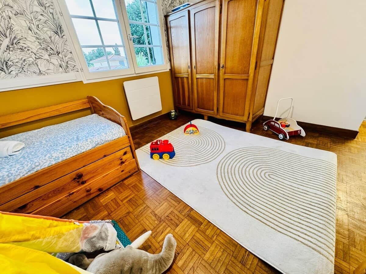 A child's room with a bed, wooden wardrobe, and toys on a patterned rug.