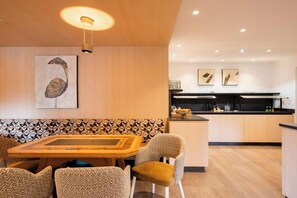 Ein modernes Interieur mit Essbereich, der einen Holztisch und stilisierte Stühle umfasst, einer Küche mit schwarzem Arbeitsplatz und einer Wand mit geometrischem Rückwandmuster.