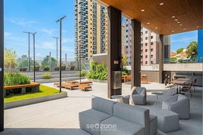 A modern outdoor seating area with sofas and tables, surrounded by greenery and a view of high-rise buildings. - Comfort in Condo. w/ Pool & Balcony Lss0705 (São Paulo)