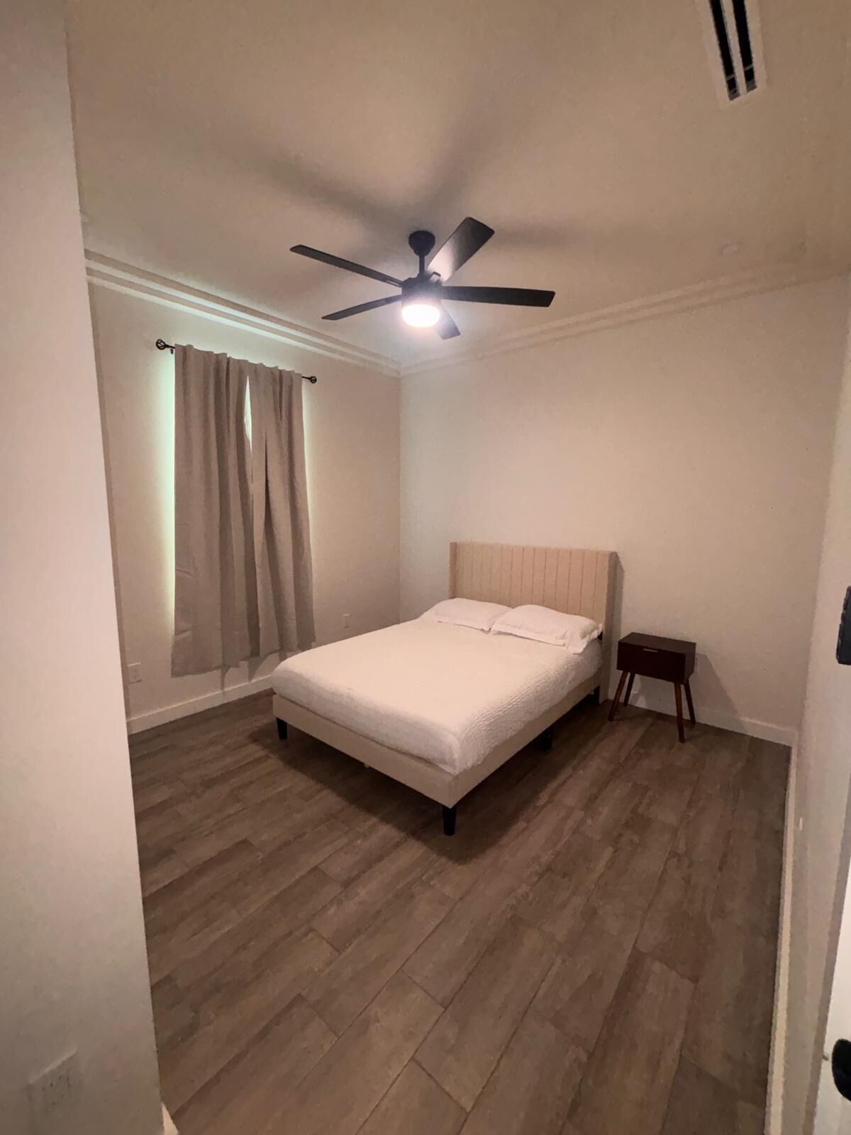 A bedroom with a bed, a ceiling fan, a nightstand, and a window with curtains.