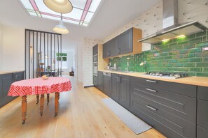 A modern kitchen with dark cabinets, a green backsplash, and a wooden dining table with a checkered tablecloth. - La Mouvalloise - Maison de charme avec jardin (Mouvaux)