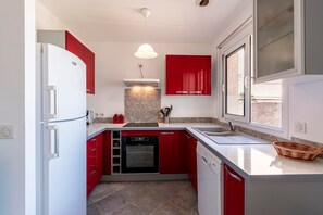 A compact kitchen with red cabinets, a white refrigerator, a built-in oven, and a window providing natural light. - A Casa di Porticcio - Avec terrasse vue mer (Grosseto-Prugna)