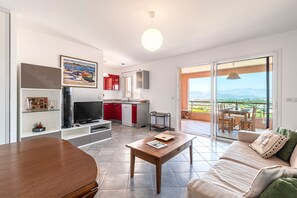 A modern living room with a TV, a wooden coffee table, and a sofa. There's a kitchen area with red cabinets and a dining area with a table and chairs. The room has a large sliding glass door leading to a balcony with a view of the mountains. - A Casa di Porticcio - Avec terrasse vue mer (Grosseto-Prugna)