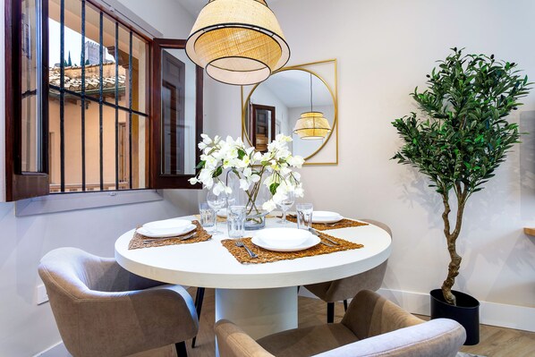 A round dining table with white plates, clear glasses, and a vase of white flowers. There are two chairs with a beige upholstery. A large mirror hangs on the wall, and a potted plant is visible to the right. - Genteel Home Santa Ines (Granada)