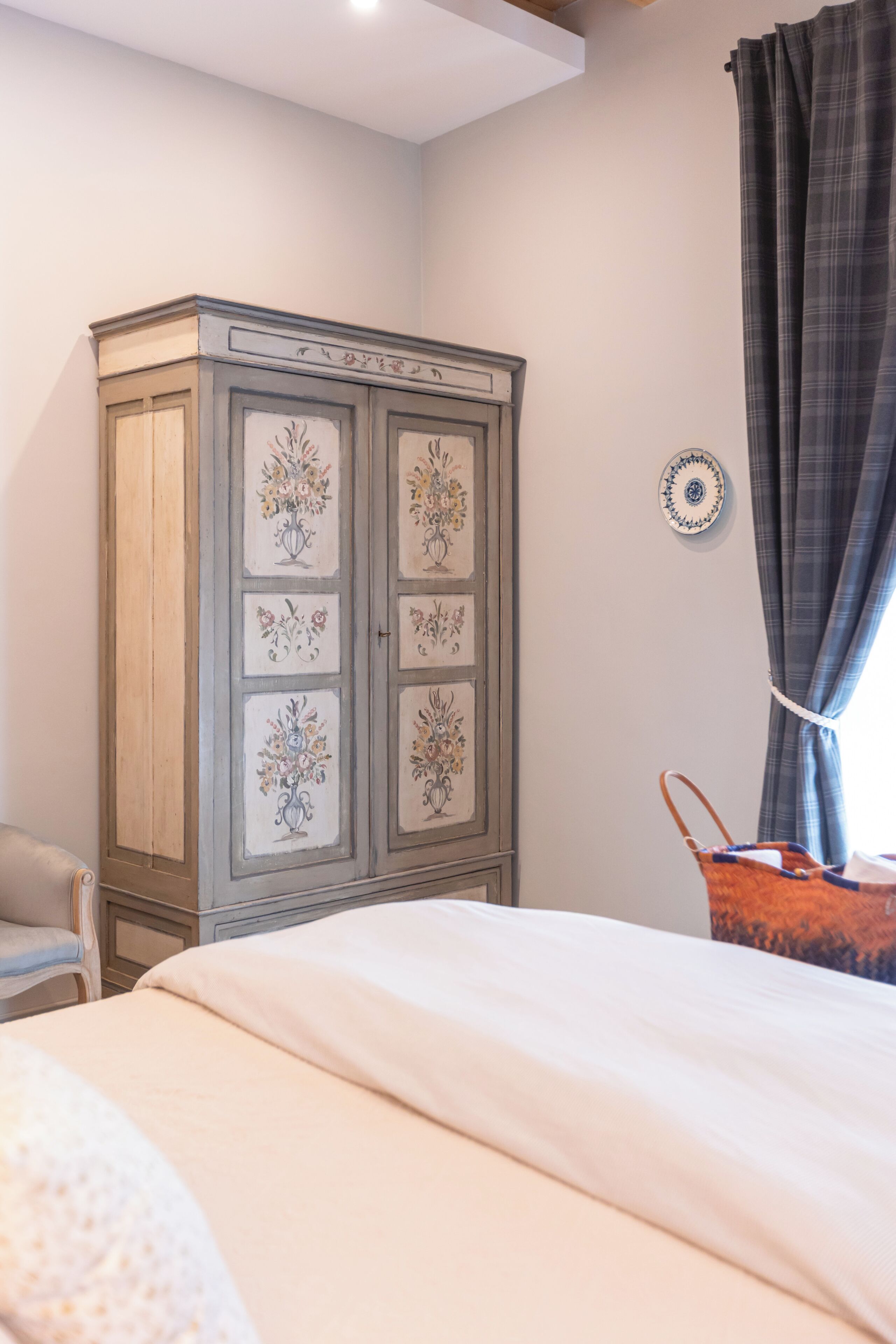 A bedroom with a bed, a wardrobe with floral designs, a round wall plate, and a plaid curtain.