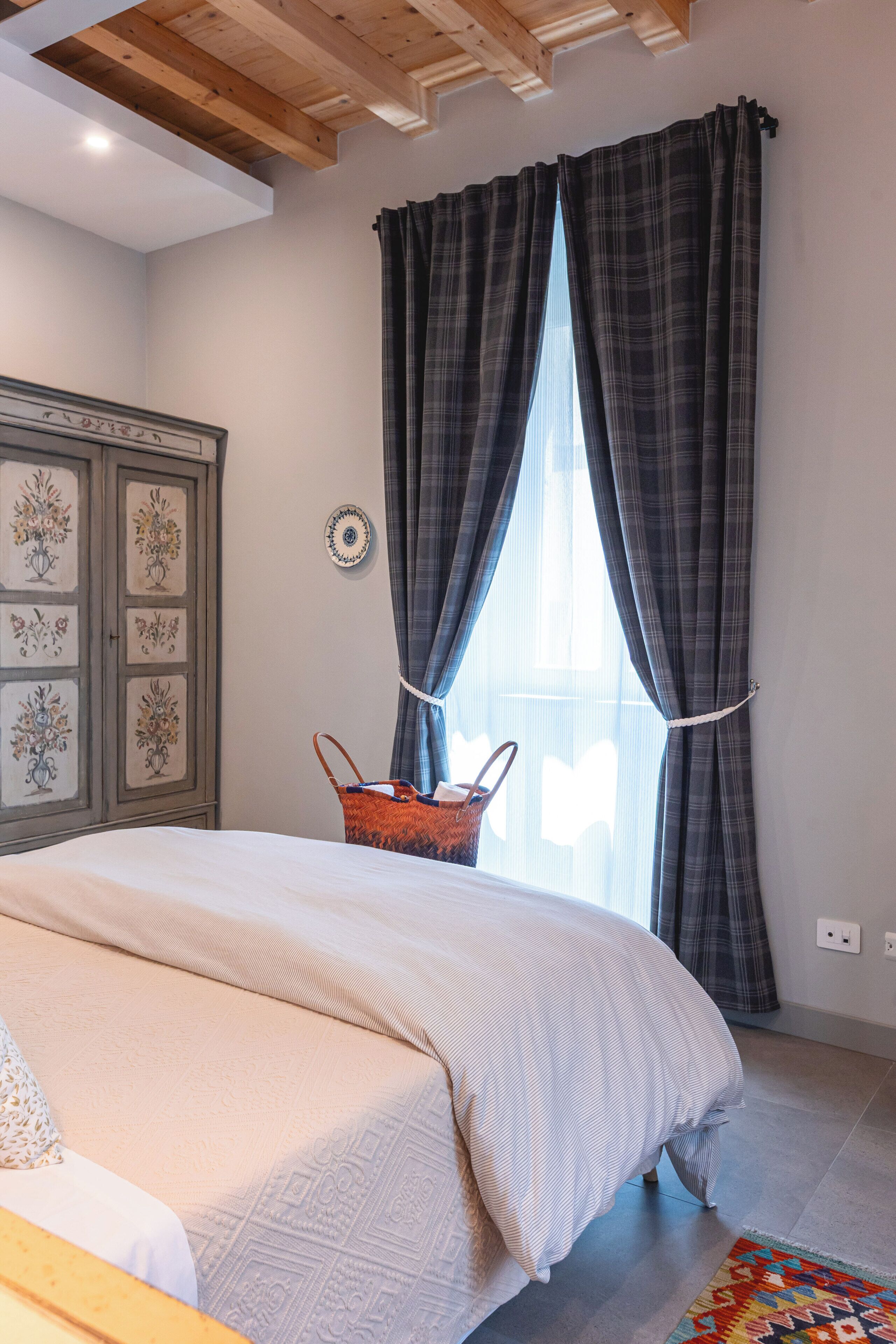 A bedroom with a bed, a wooden ceiling, a window with curtains, a wall-mounted clock, and a decorative cabinet.