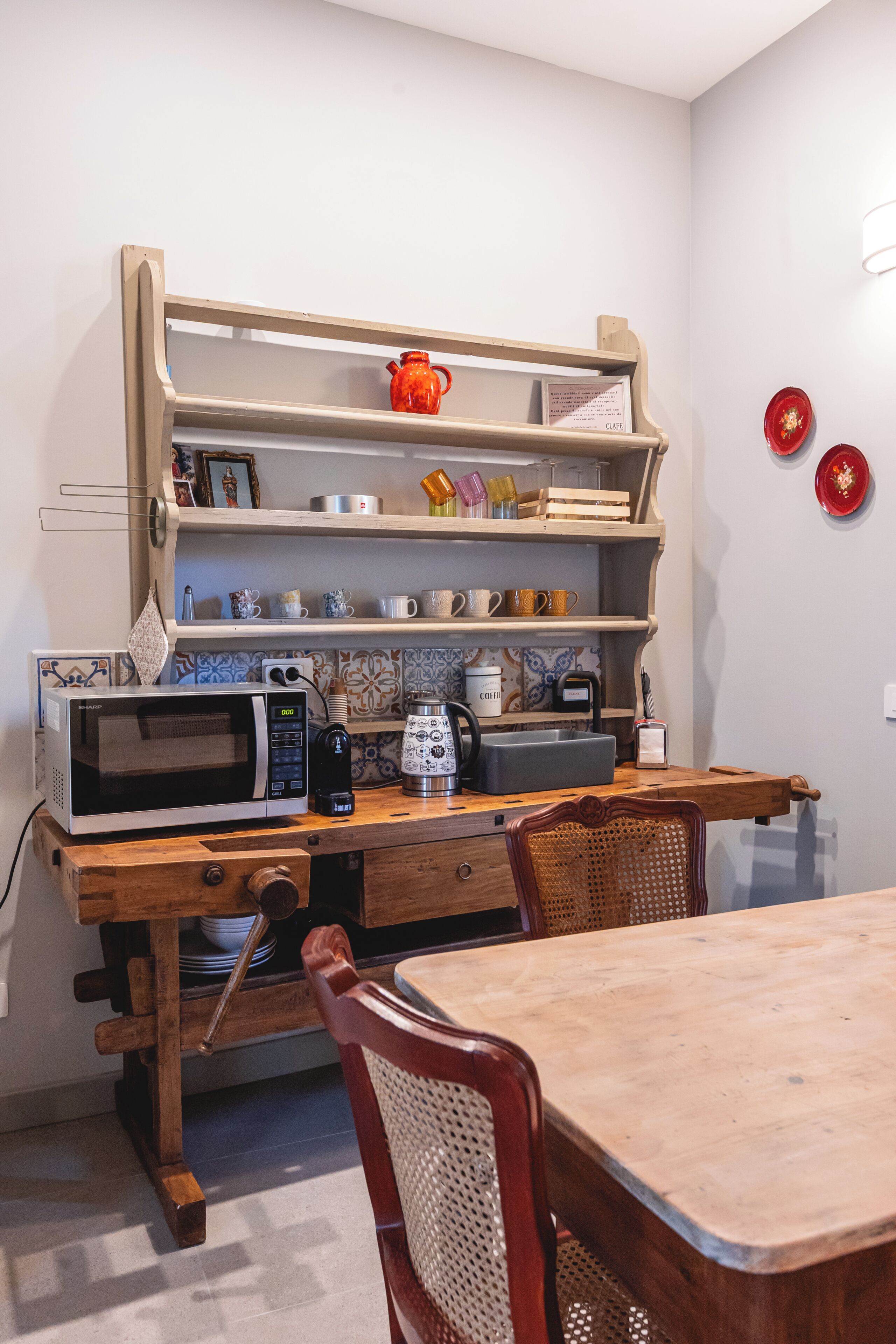 Eine Küche mit einem Holztisch, Stühlen, einer Mikrowelle, einer Kaffeemaschine und einem Regal mit diversen Gegenständen.